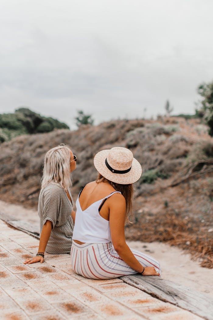 Two women sitting together outdoors enjoying a relaxed summer day in nature, wearing casual attire and hats.